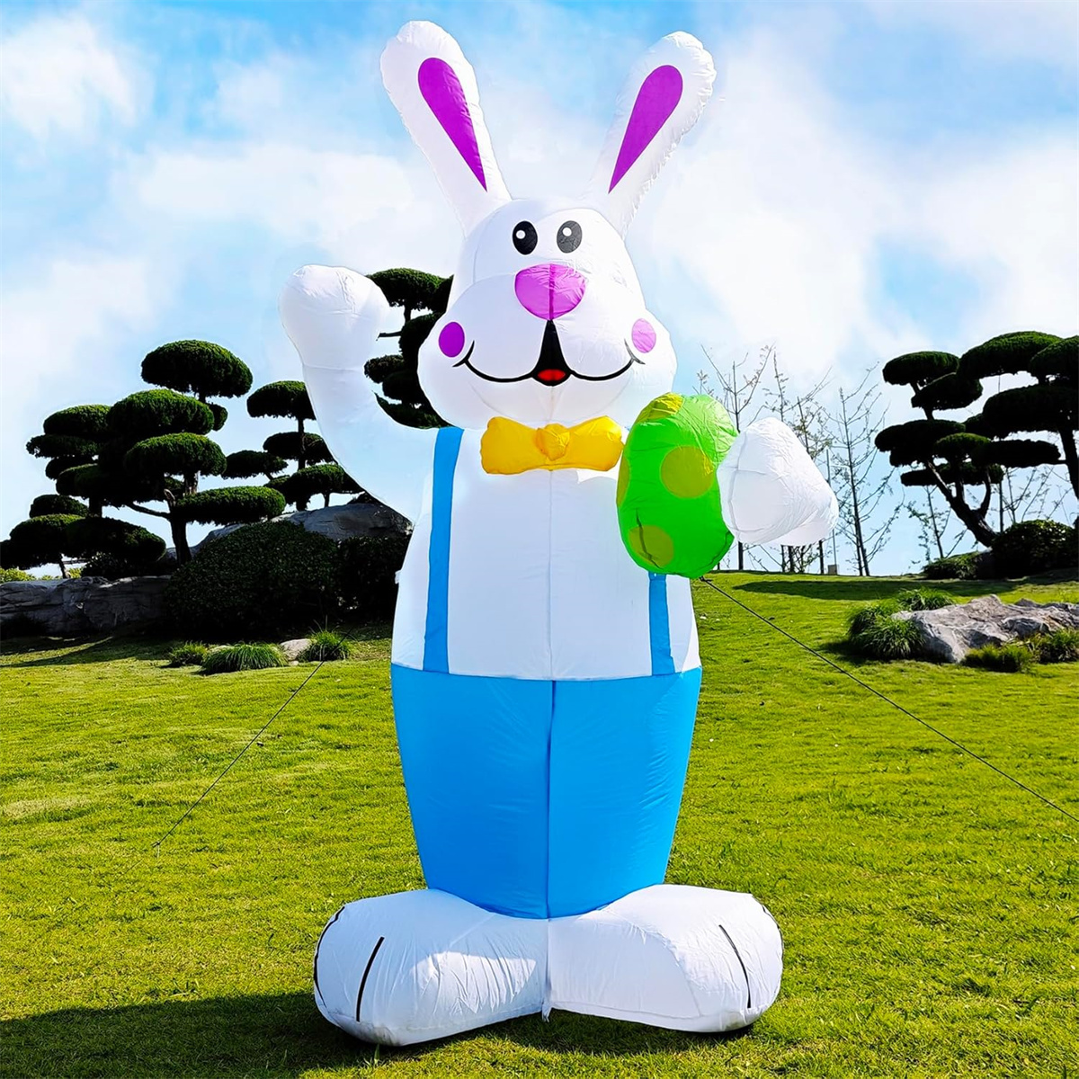 Ein großes aufblasbares Osterkaninchen, das blaue Latzhosen trägt und ein grünes Ei hält, auf grünem Gras.
