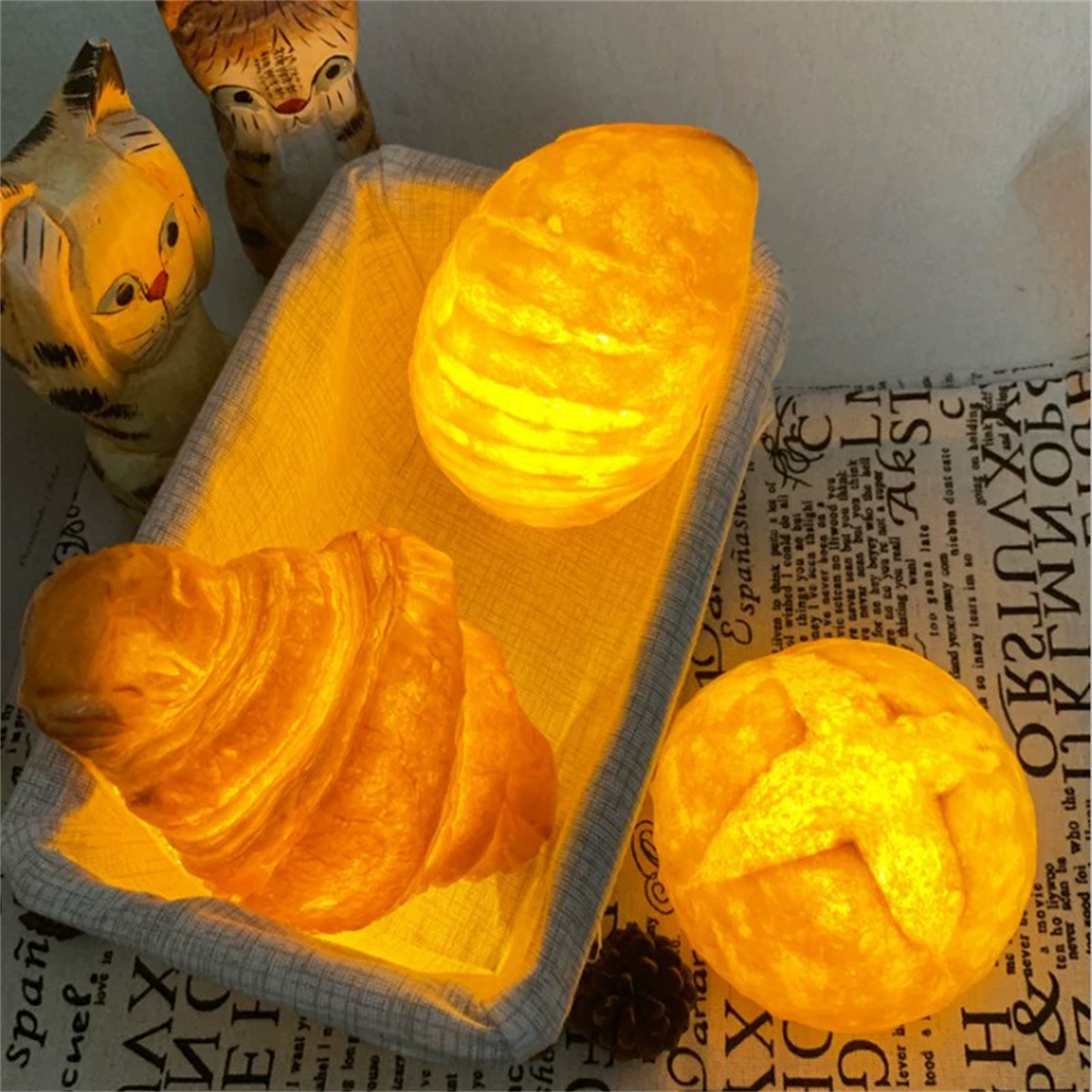 Ein Korb mit beleuchtetem Brot. Ein Croissant, rundes Brot und ein weiteres Brot sind darin. Hinten sind zwei Holzkatzenfiguren.