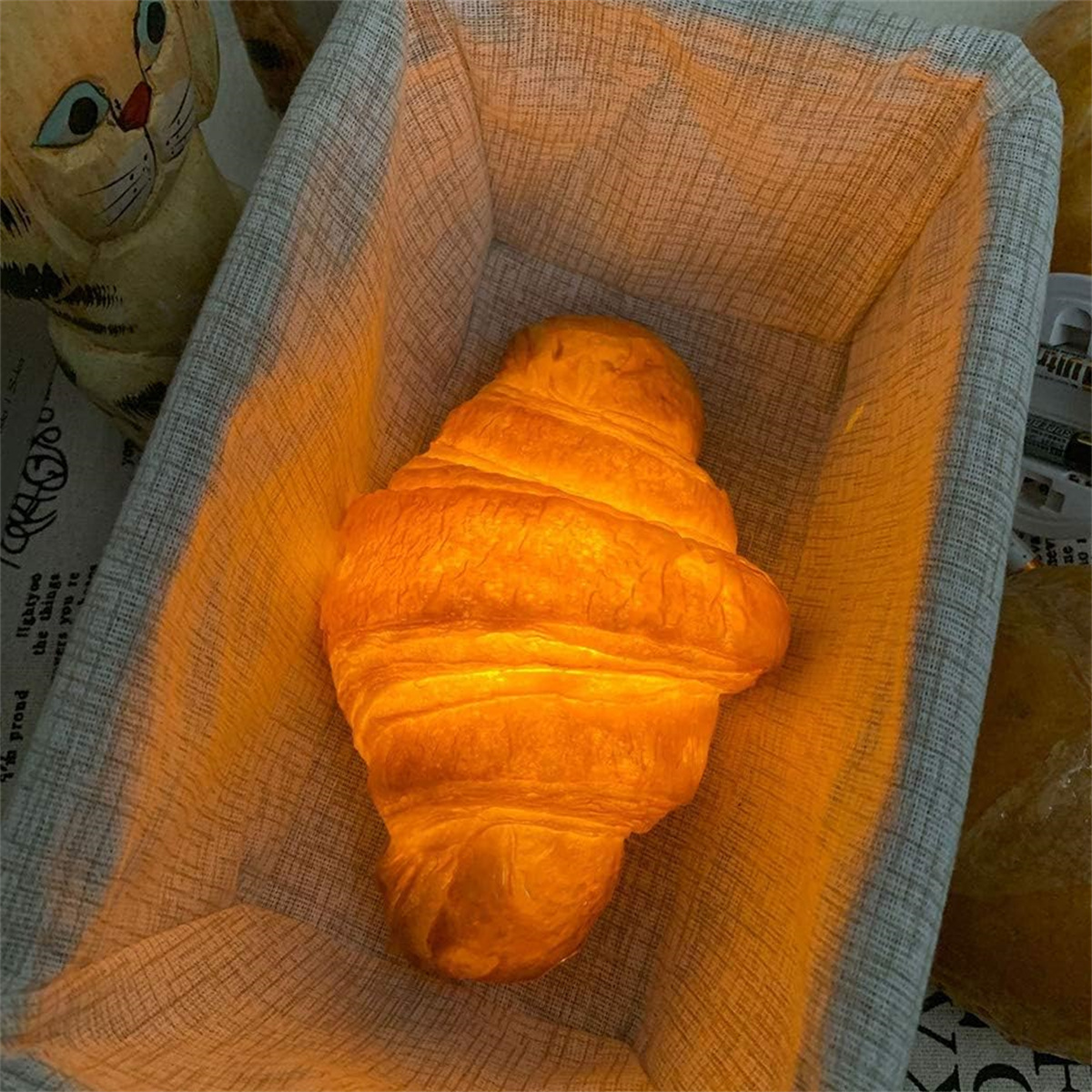 Ein goldbraunes Croissant in einem mit Stoff ausgekleideten Korb. Eine Katzenfigur im unscharfen Hintergrund.