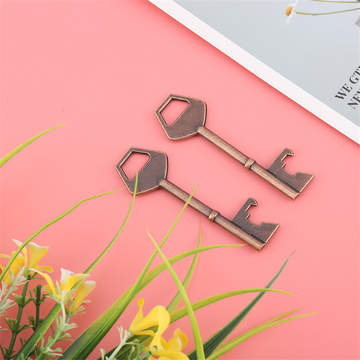 Zwei bronzefarbene Schlüssel mit Flaschenöffner-Design, auf pinkem Hintergrund mit Blumen.