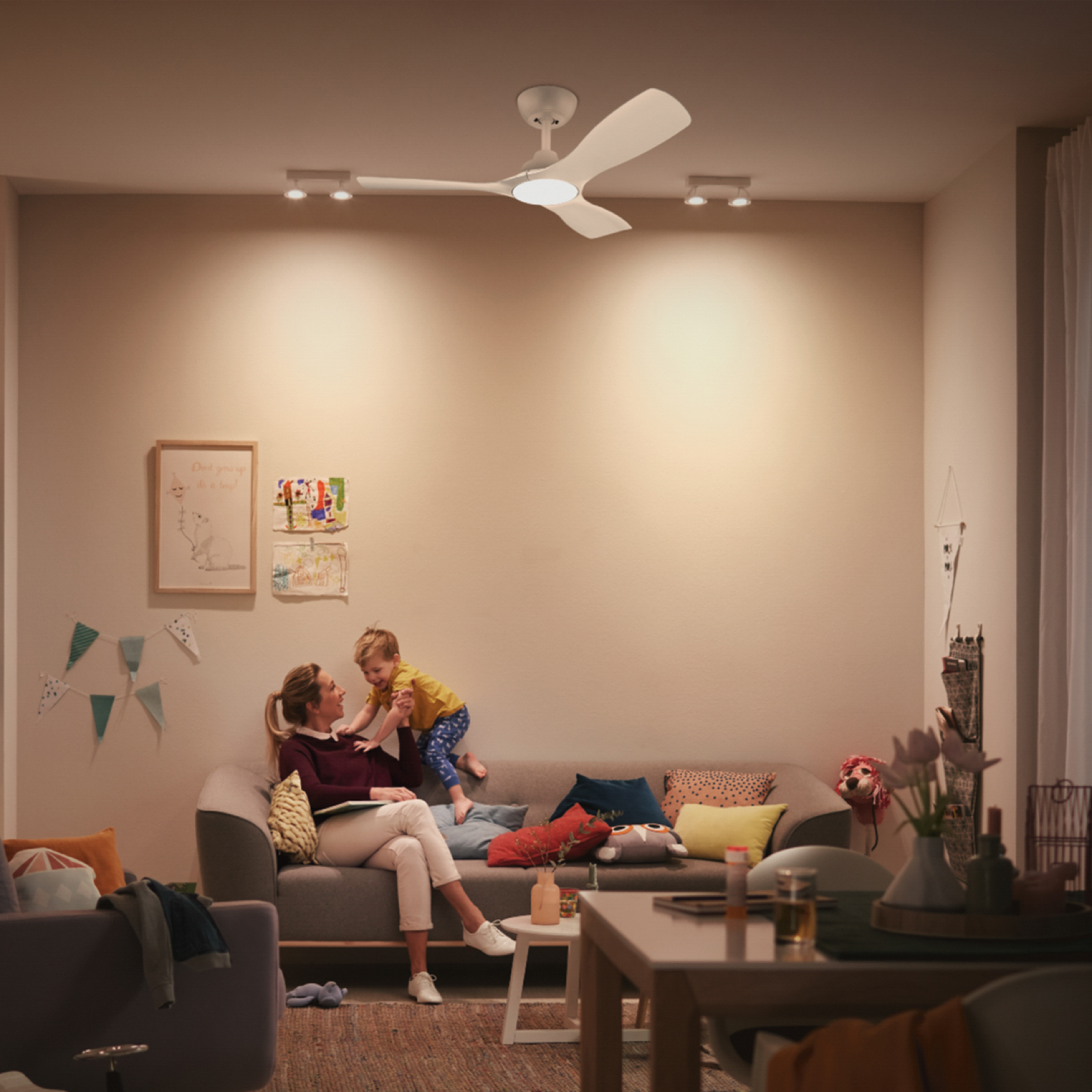 Sala de estar con mujer, niño, sofá, ventilador y luces. Paredes beige.
