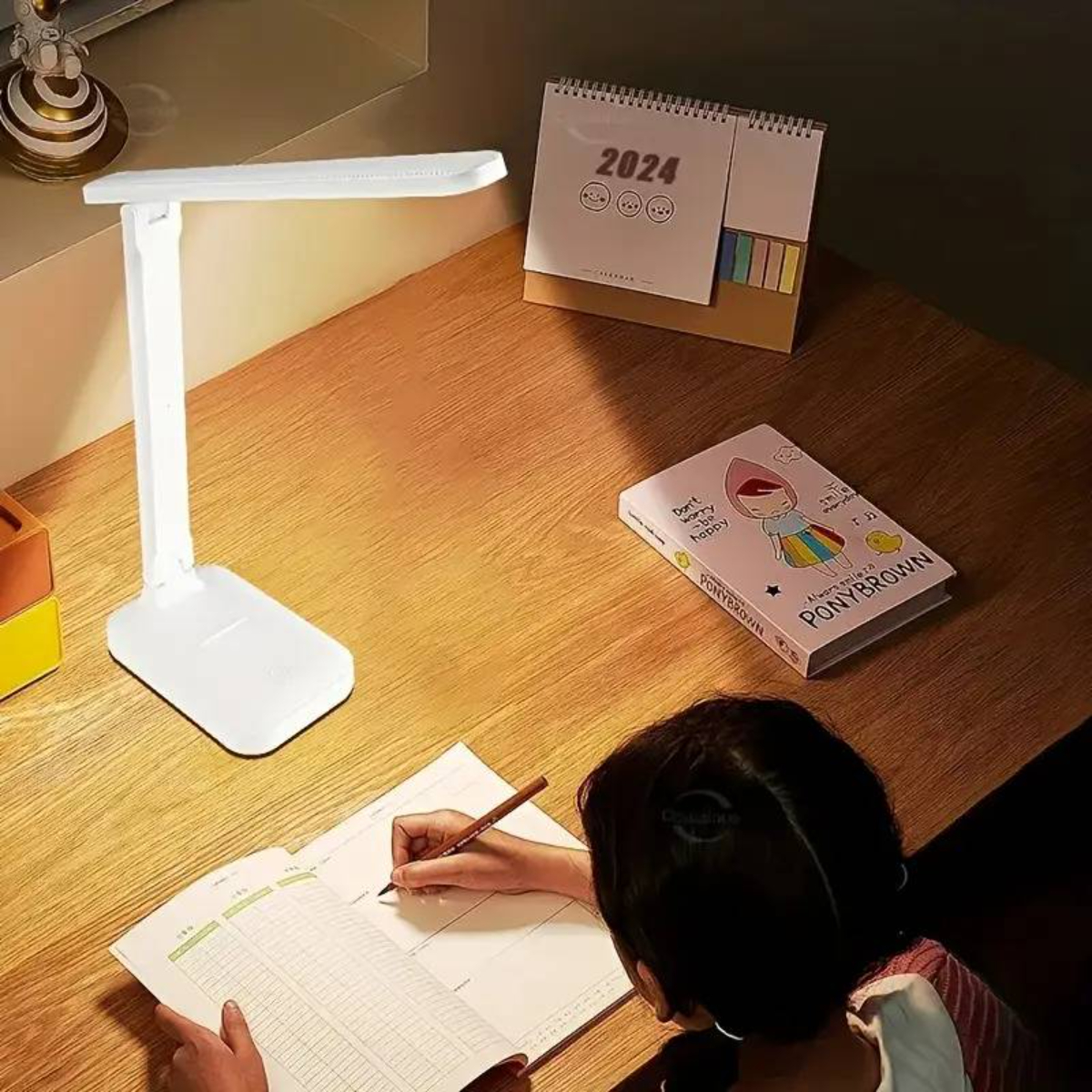 Ein Schreibtisch mit weißer Lampe, Kalender, Buch und einer Person, die schreibt. Holzoberfläche, warme Beleuchtung.