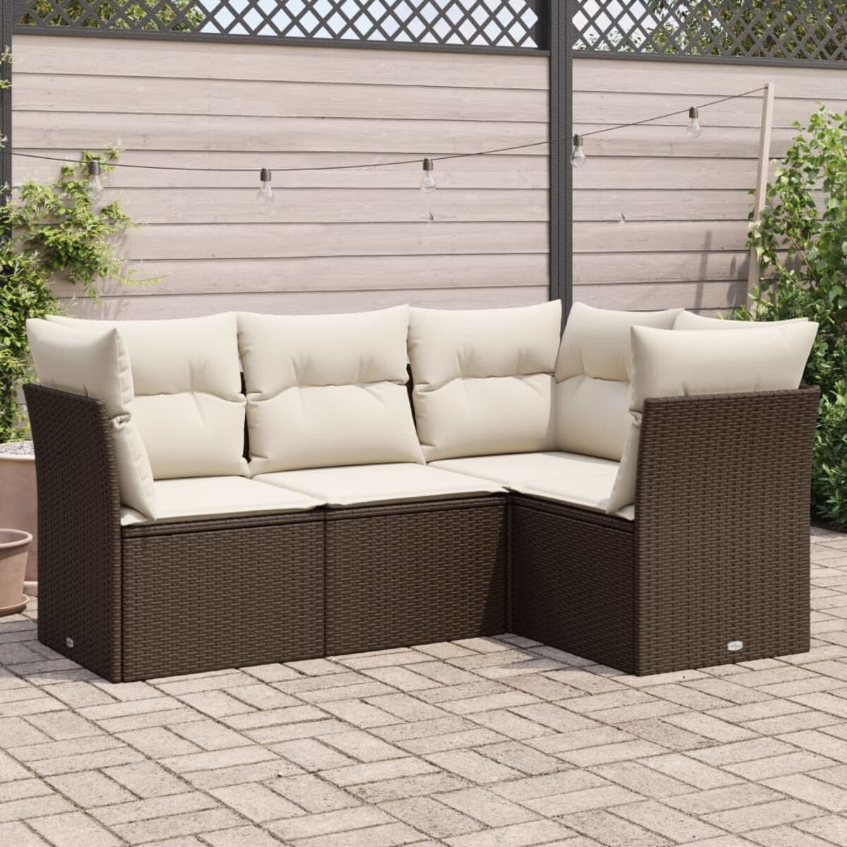 Un divano componibile da esterno in vimini marrone con cuscini color crema, su un patio in mattoni, con una recinzione in legno.