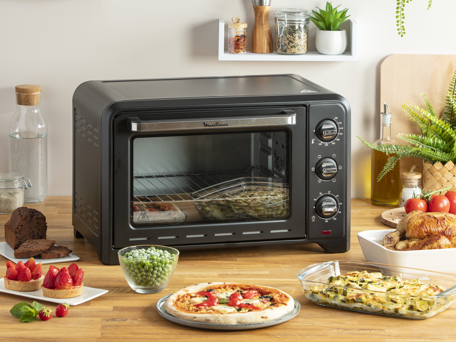 Czarny piekarnik Moulinex z zawartością, w tym pizzą i pieczonym jedzeniem na drewnianym stole.