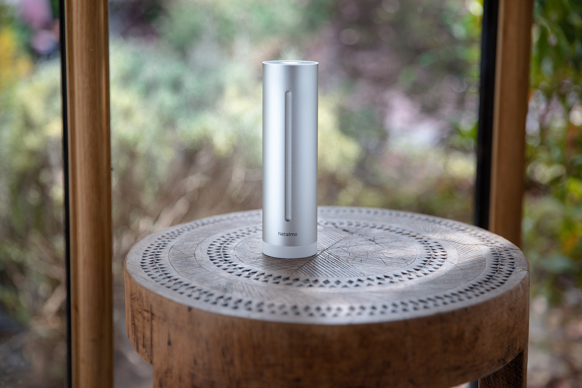 Srebrne, cylindryczne urządzenie Netatmo na drewnianym stole z rozmytym tłem okna.