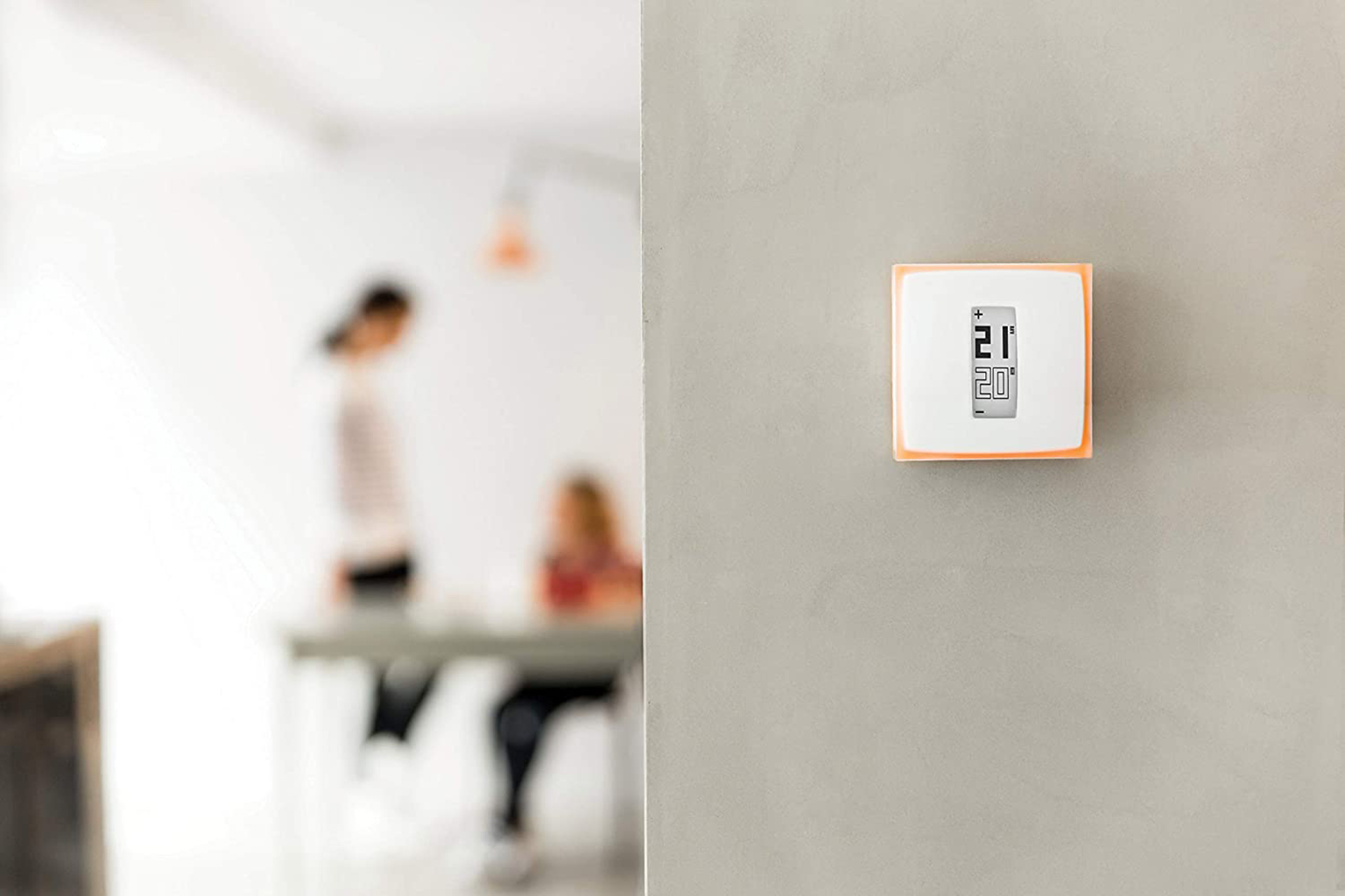 Thermostat mounted on a gray wall. Shows 21 degrees. Blurred background with people in room.