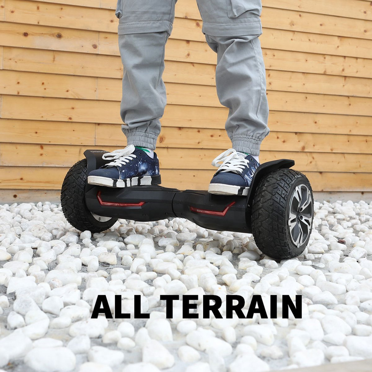 A person standing on a black and red hoverboard on white stones, with wooden planks in the background.