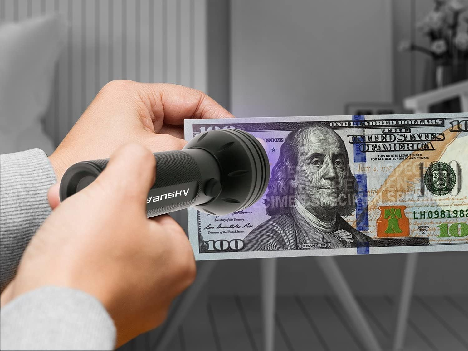 Hand hält Taschenlampe über einen 100-Dollar-Schein. Die Rechnung ist teilweise beleuchtet, wodurch Details sichtbar werden.