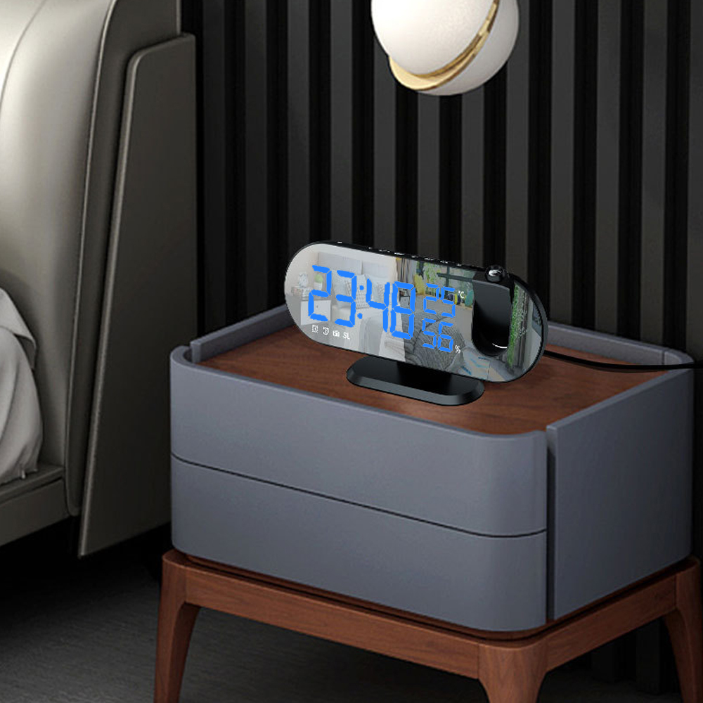 Ein schwarzer digitaler Wecker auf einem grauen Nachttisch mit einem Bett im Hintergrund.