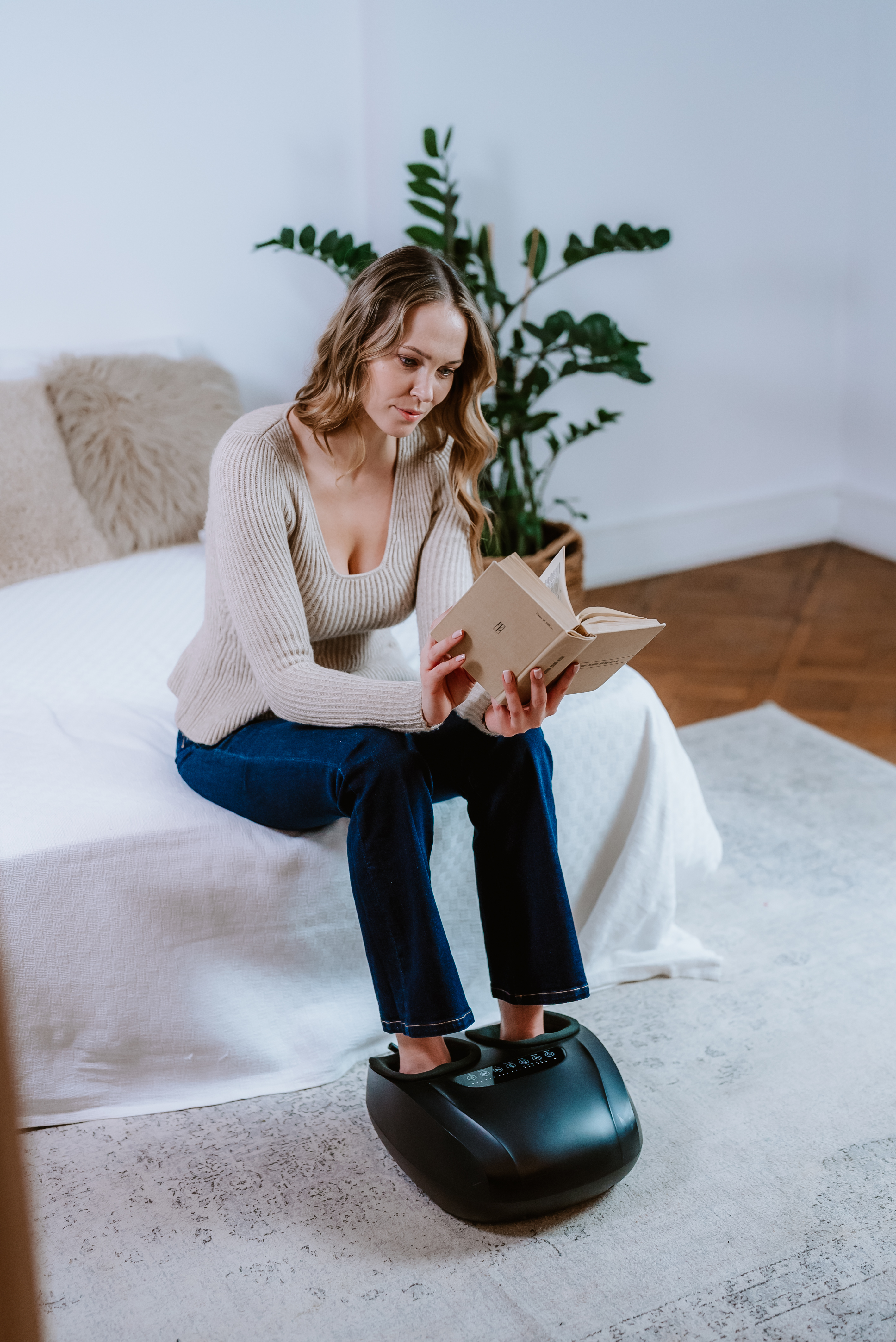 Eine Frau sitzt auf einem Bett und liest ein Buch. Ihre Füße stecken in einem schwarzen Fußmassagegerät.