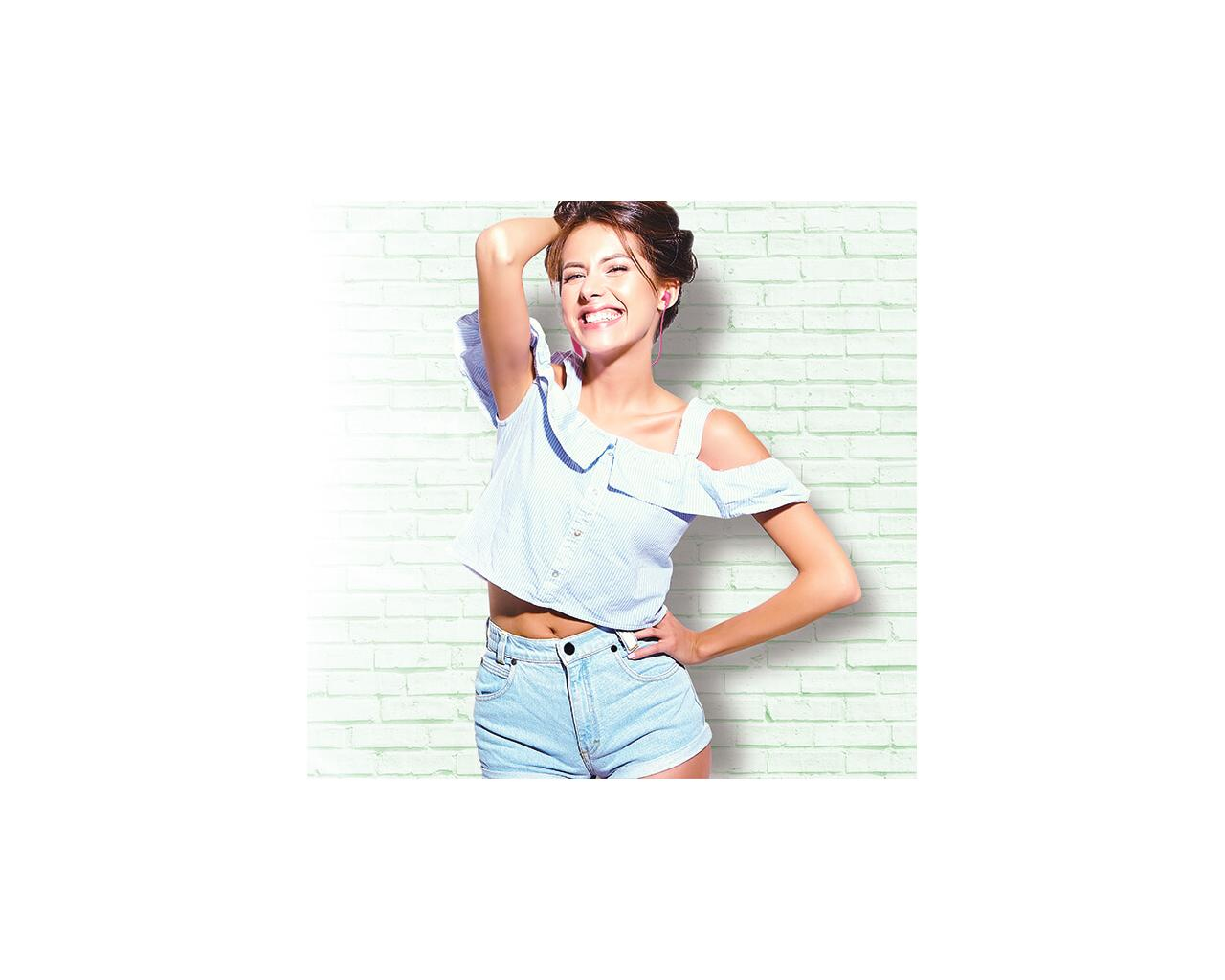 Uśmiechnięta kobieta stojąca przed zieloną ceglaną ścianą, ubrana w niebieski top i dżinsowe szorty.