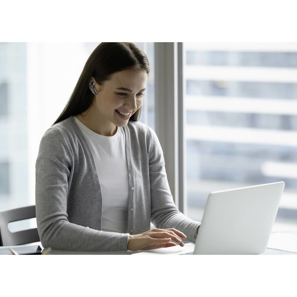 Frau mit Ohrhörern, lächelnd, arbeitet im Büro an einem Laptop. Hintergrund: Stadtbild.
