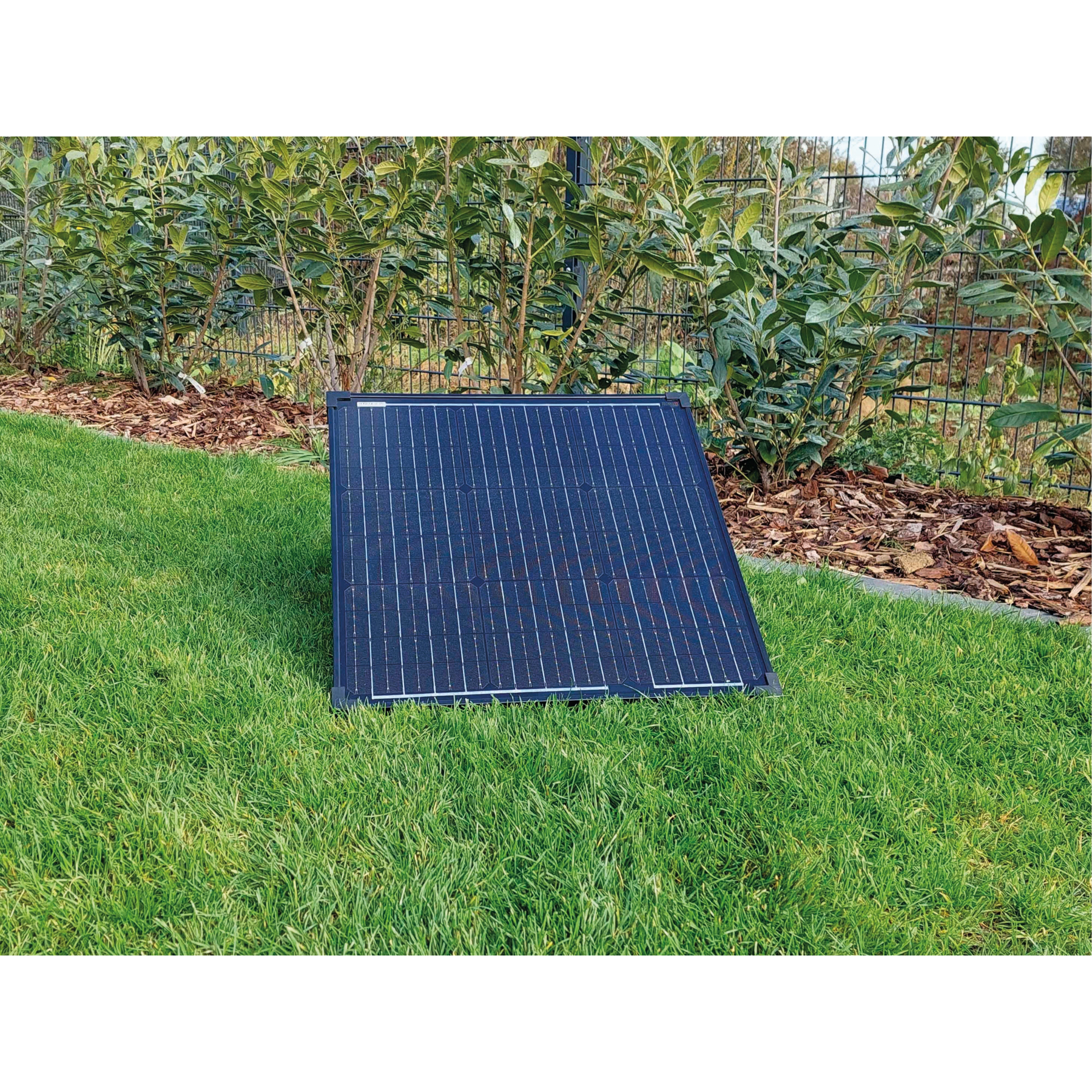 Panel słoneczny umieszczony na zielonej trawie w ogrodzie z ogrodzeniem i krzewami w tle.
