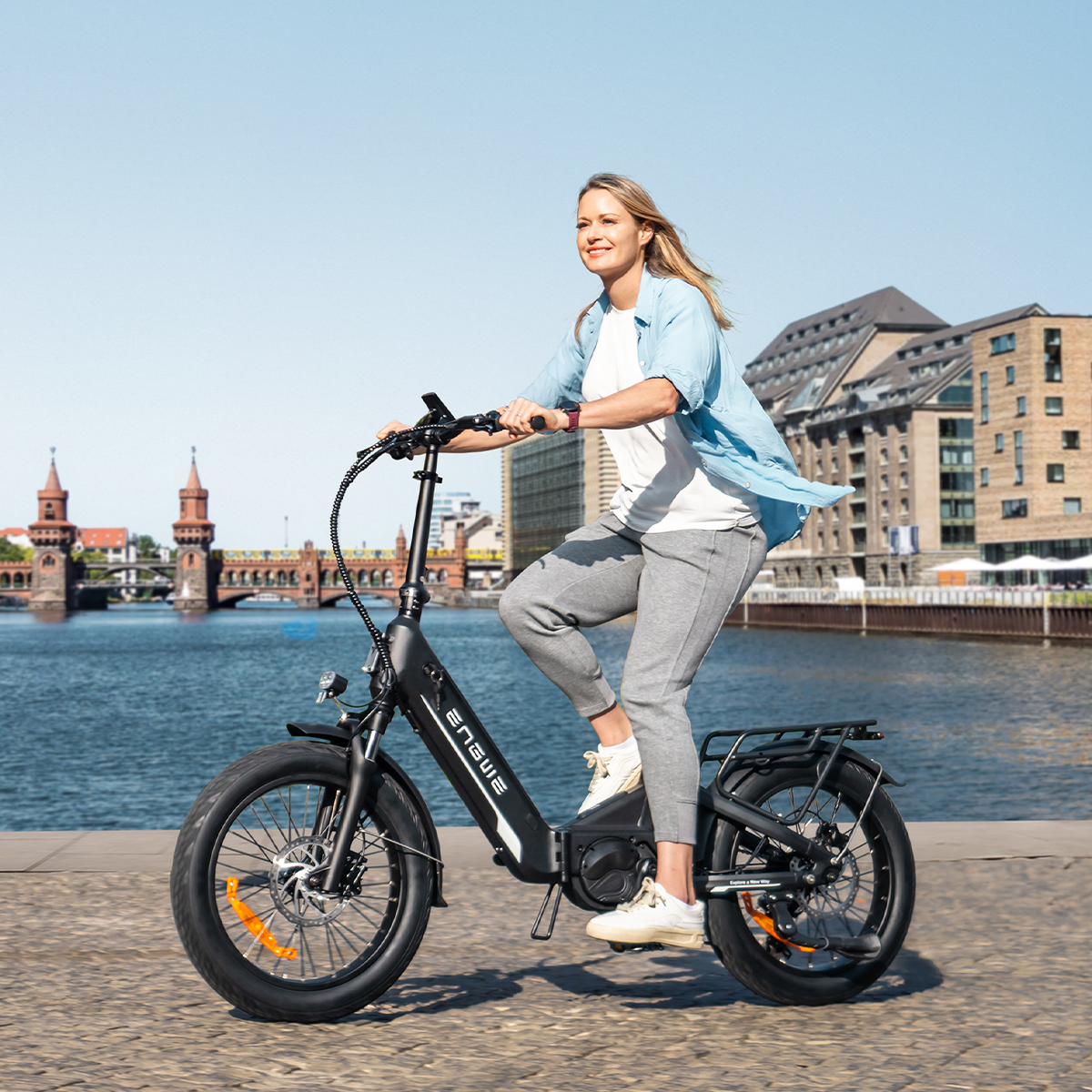 Donna che guida una bici elettrica nera in una giornata di sole. Fiume ed edifici sullo sfondo.
