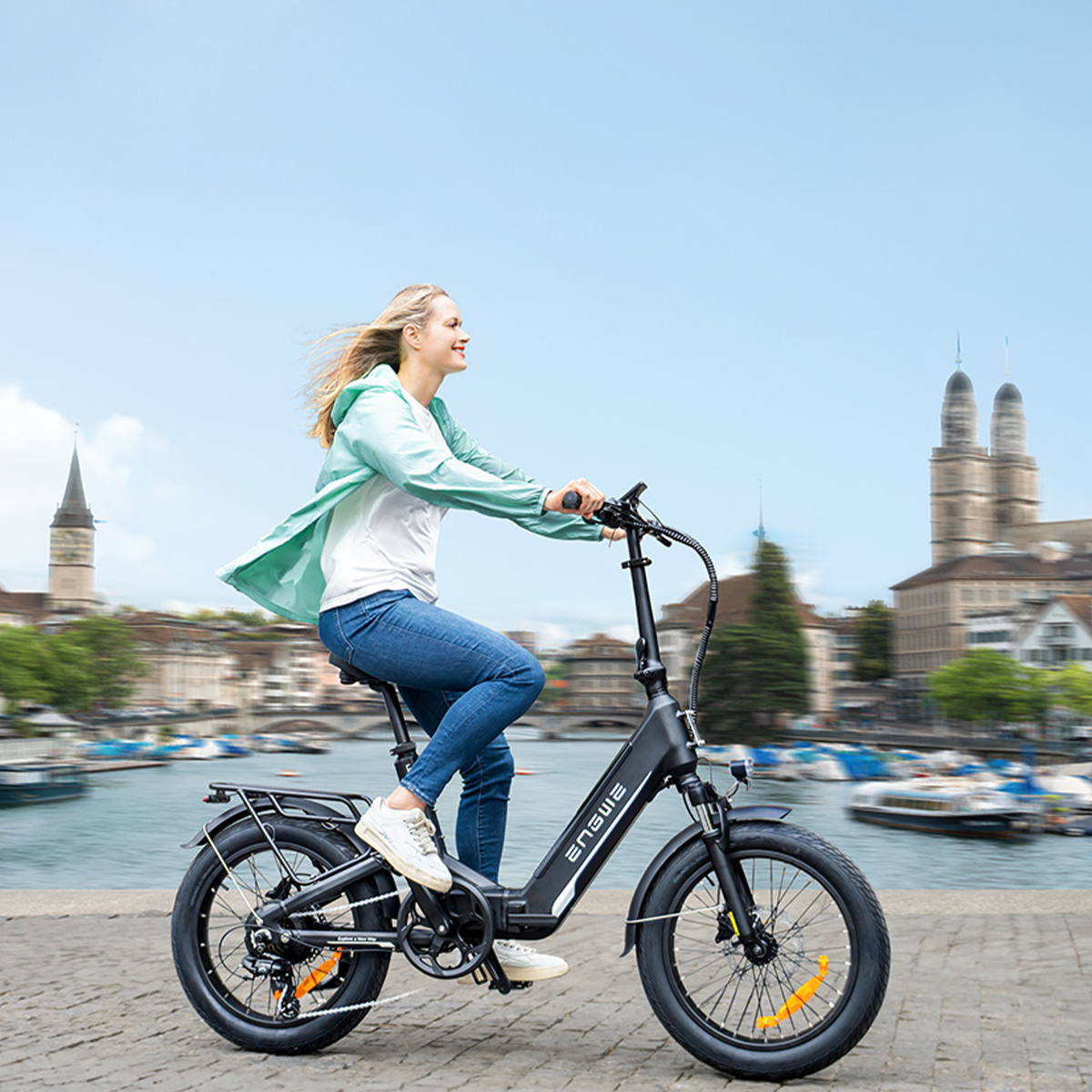 Donna in bicicletta elettrica nera vicino all'acqua, edifici sullo sfondo.