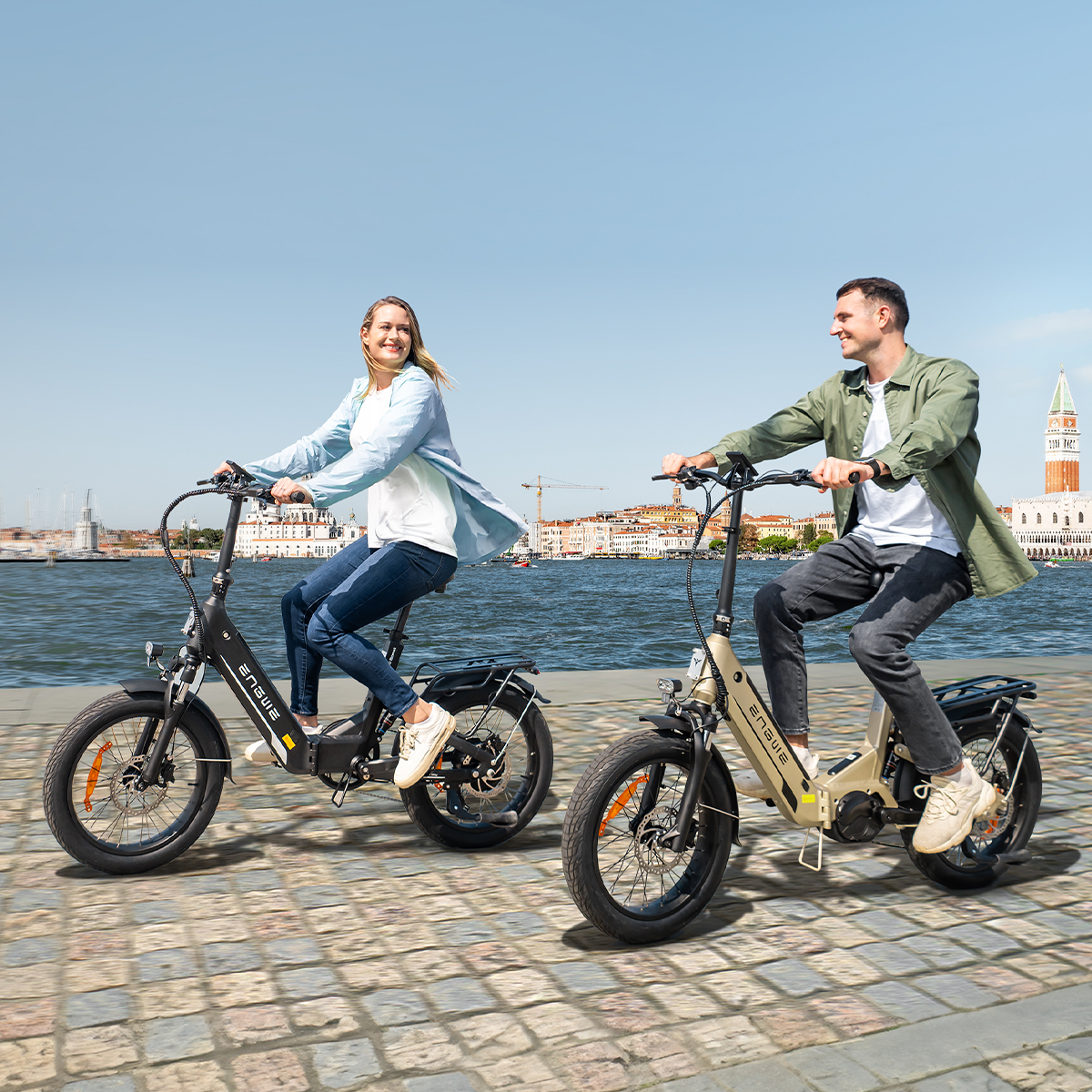 Due persone in bicicletta elettrica nera e beige lungo un lungomare con edifici.