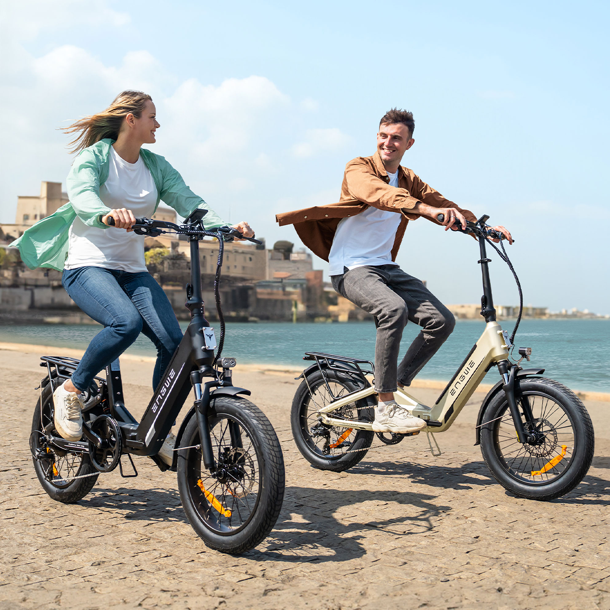 Due persone in bicicletta elettrica su un sentiero vicino all'acqua. Scena soleggiata.