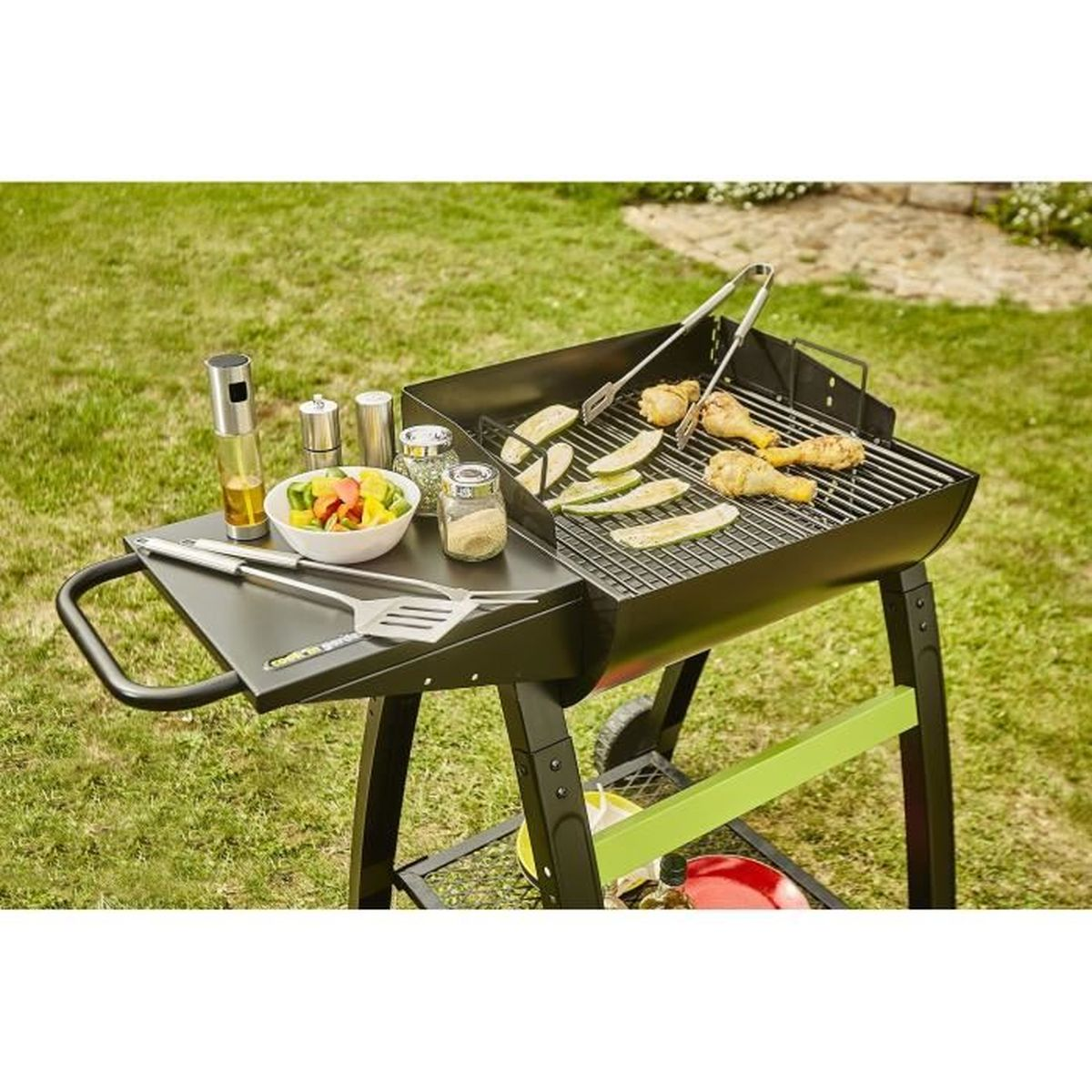 Grill mit Essen, einschließlich Hühnchen und Gemüse, auf Gras. Utensilien und Gewürze sind ebenfalls vorhanden.