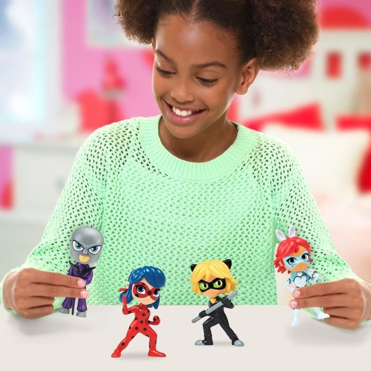 Ein lächelndes Mädchen, das vier Miraculous Ladybug-Figuren hält. Weiße Oberfläche mit rosa Hintergrund.