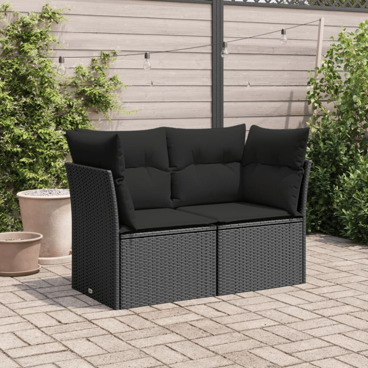 Schwarzes Outdoor-Sofa aus Korbgeflecht mit schwarzen Kissen, auf einer Ziegelterrasse mit Pflanzen.