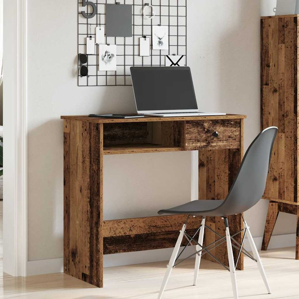 Holztisch mit Laptop, Stuhl und Gitter an der Wand. Rechts steht ein Schrank.