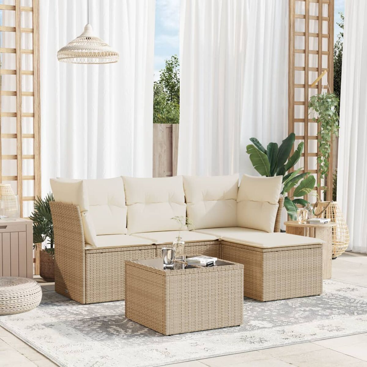 Divano componibile da esterno in vimini beige con cuscini, tavolino e tappeto, con tende bianche.