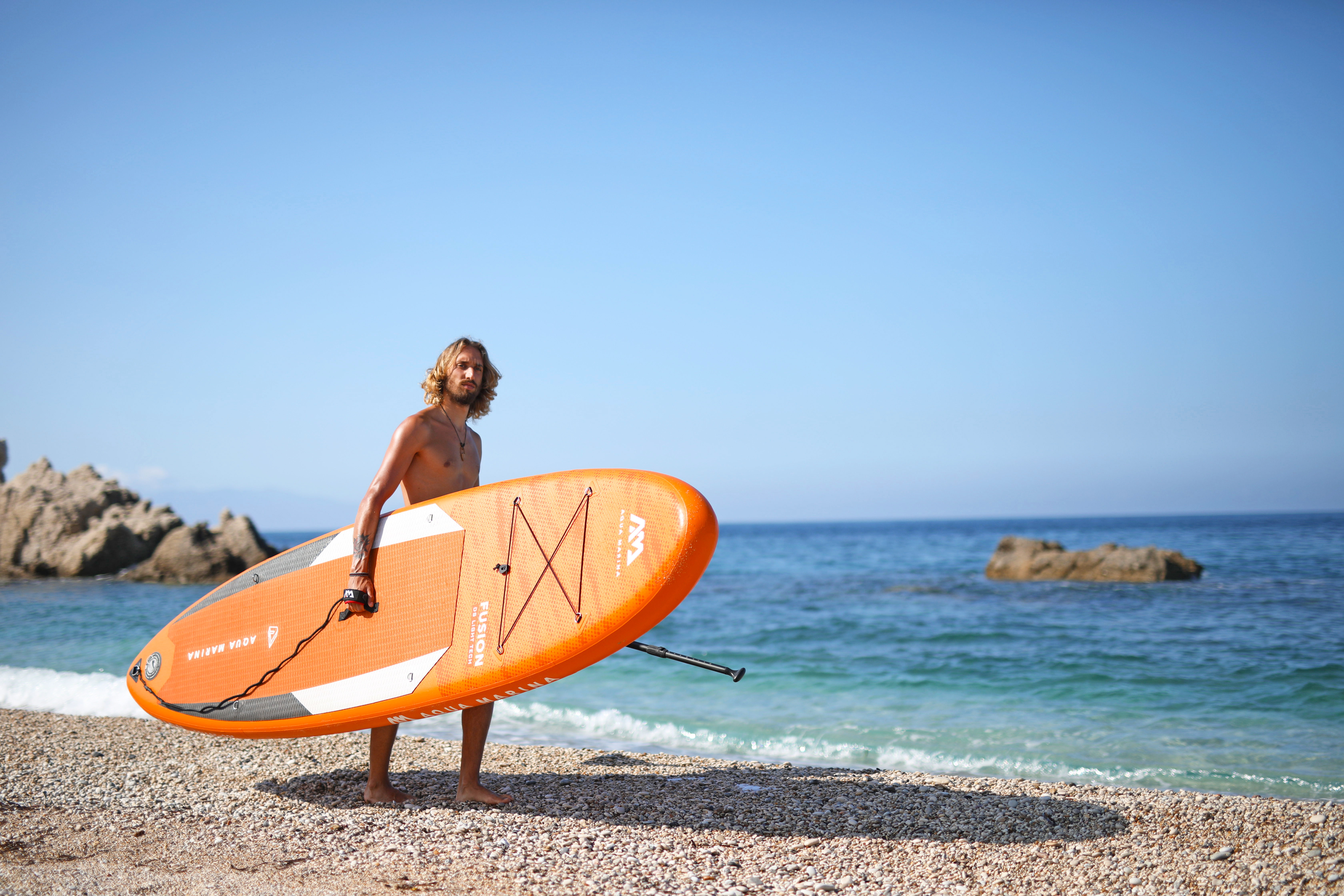 Ein Mann, der ein orangefarbenes und weißes Paddleboard an einem Strand mit blauem Wasser und Himmel hält.