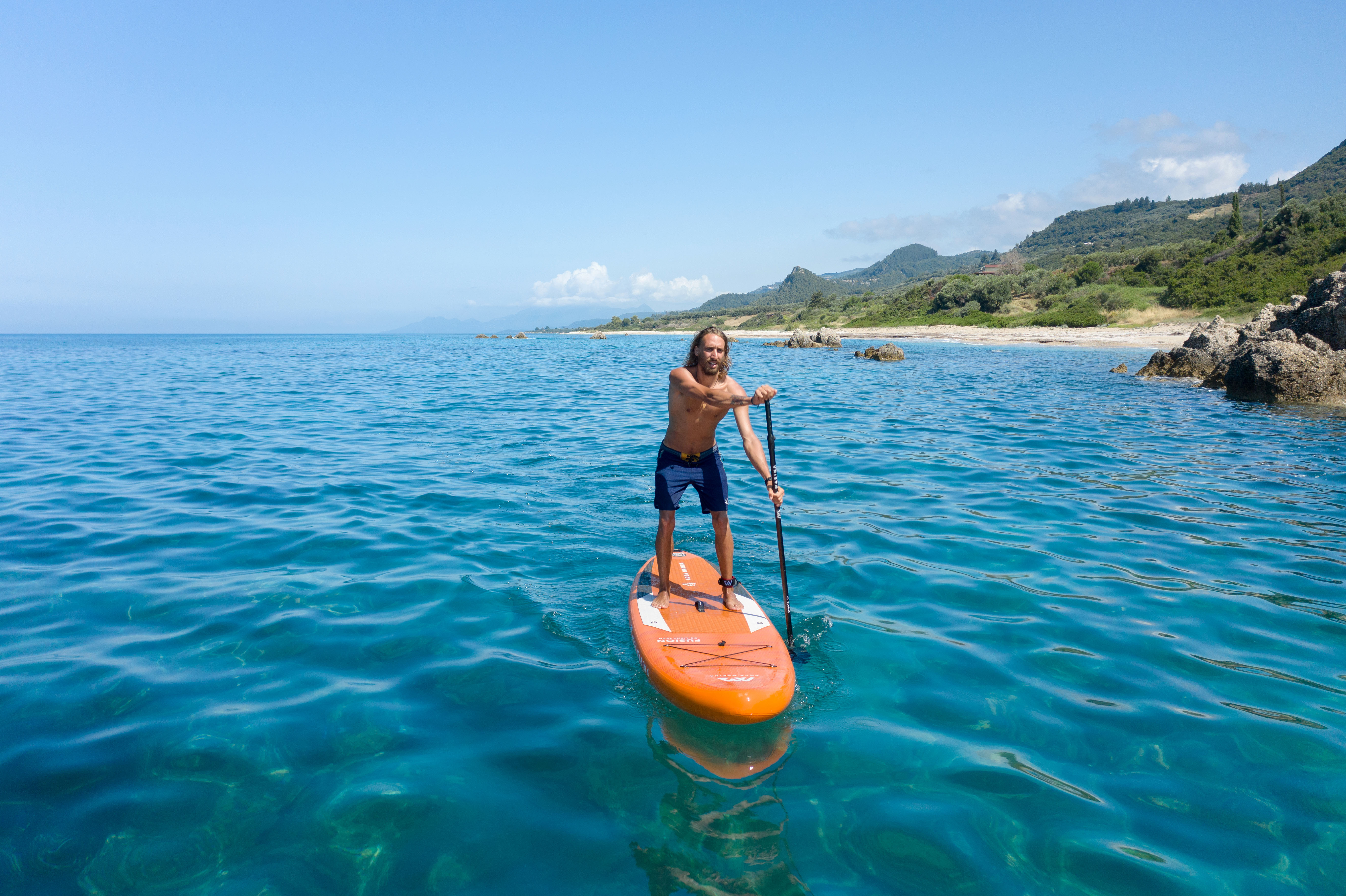 Ein Mann auf einem orangefarbenen Paddleboard auf dem Meer. Der Himmel ist blau und das Wasser klar.