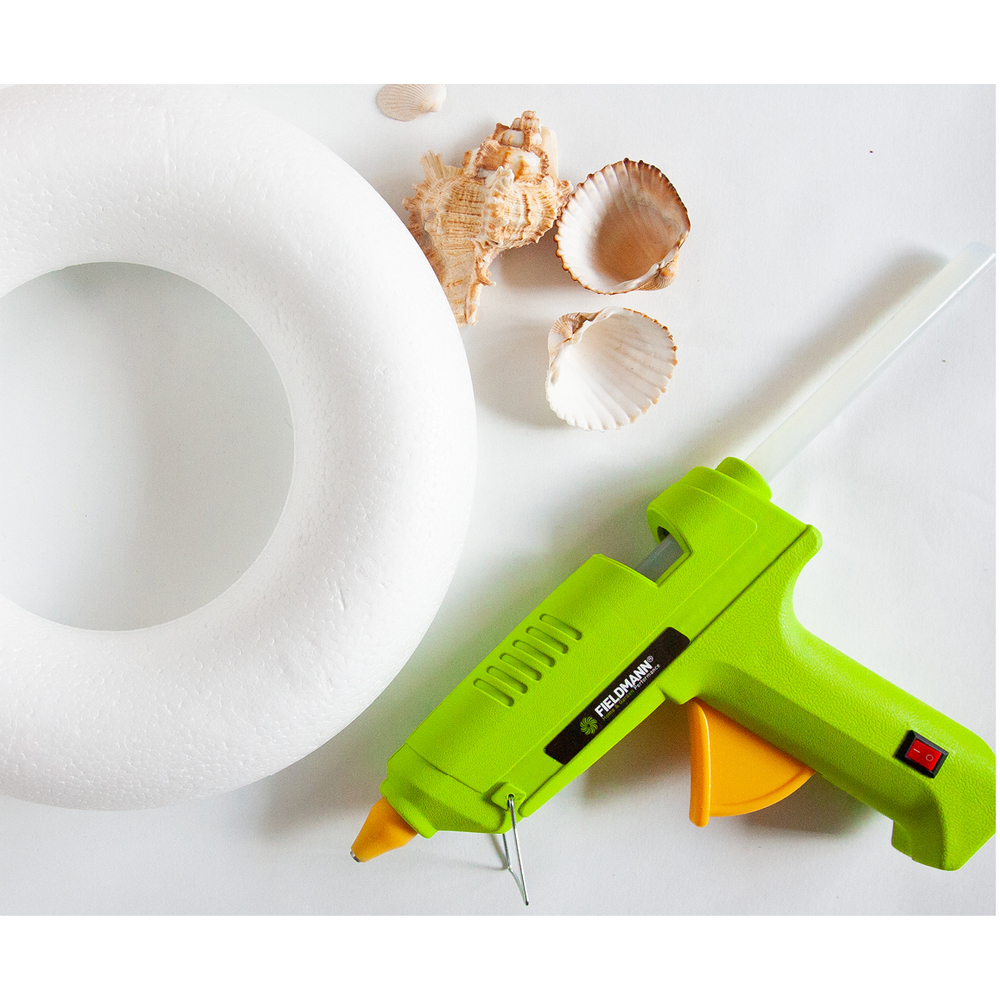 Pistola per colla verde con una ghirlanda di schiuma bianca e conchiglie su una superficie bianca.