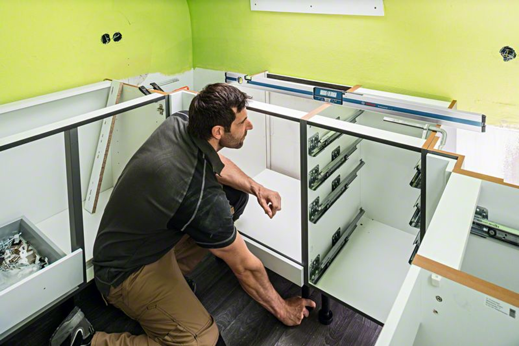 Mężczyzna w szarej koszuli montuje szafki kuchenne. Szafki są białe, a ściany zielone i niedokończone.