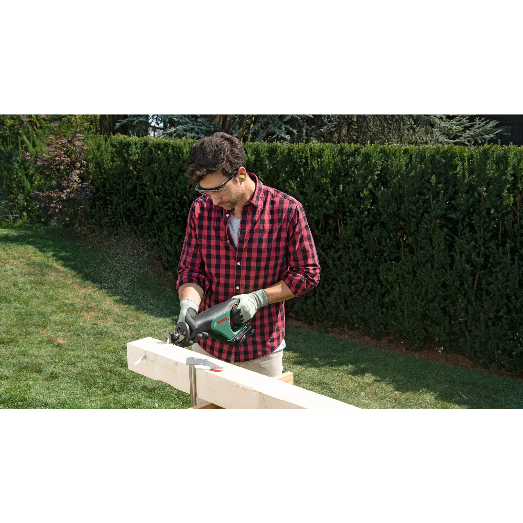 Mann sägt Holz mit einer Säbelsäge. Grüner Rasen und Hecke im Hintergrund.