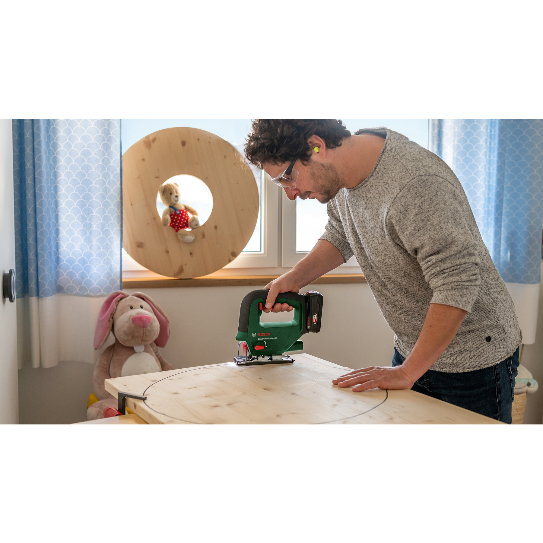 Mężczyzna używa zielonej wyrzynarki do cięcia drewnianego koła, z misiem i królikiem w tle.