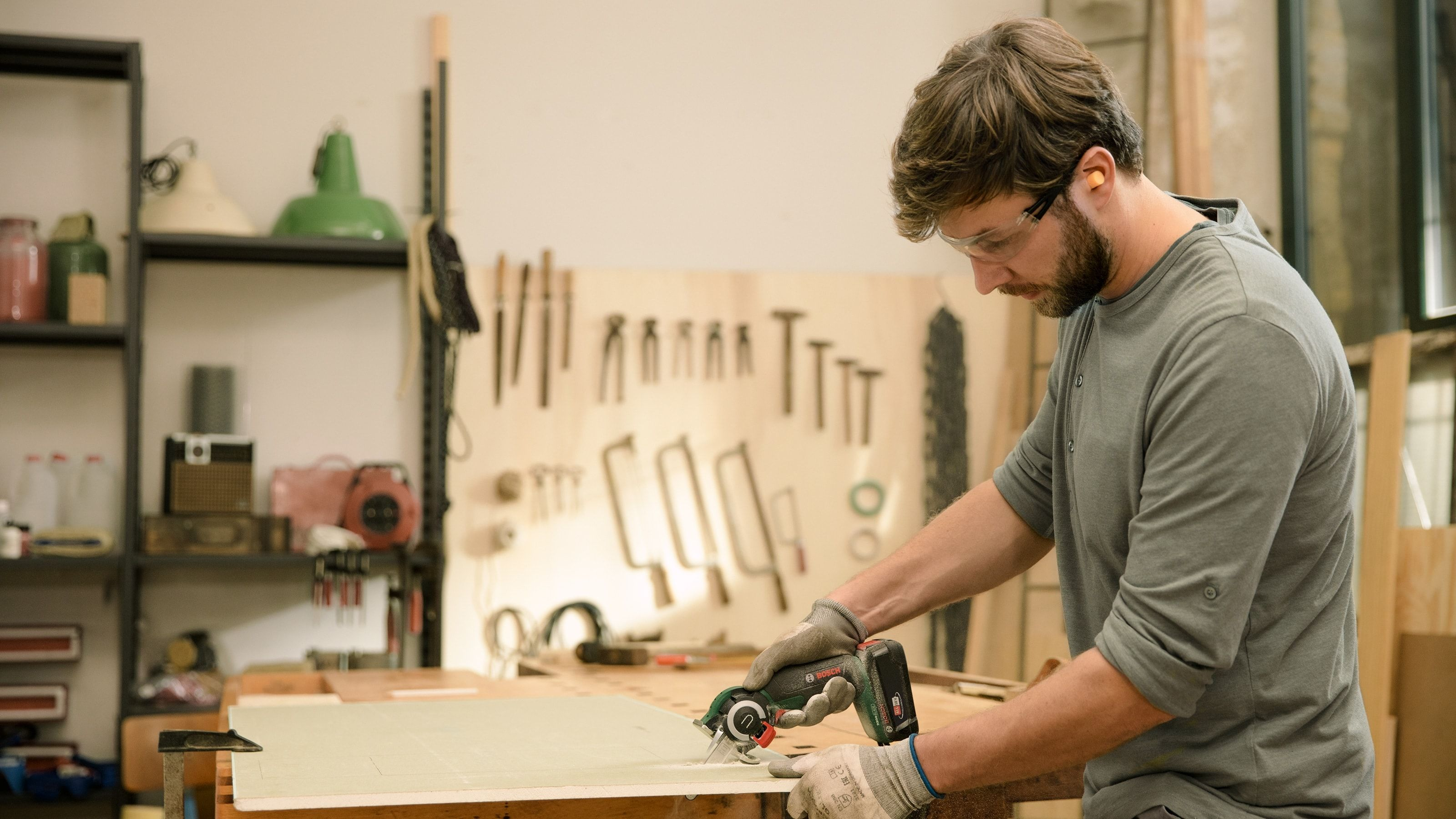 Mann sägt eine Platte in der Werkstatt. Werkzeuge und Regale sind im Hintergrund. Der Mann trägt eine Schutzbrille.