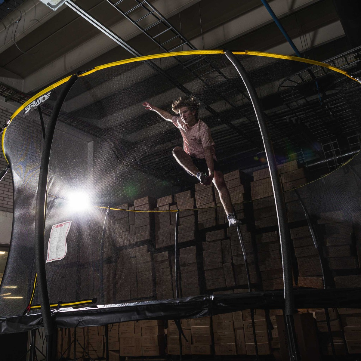 Osoba skacząca na trampolinie. Osoba jest w powietrzu. Tłem jest magazyn.