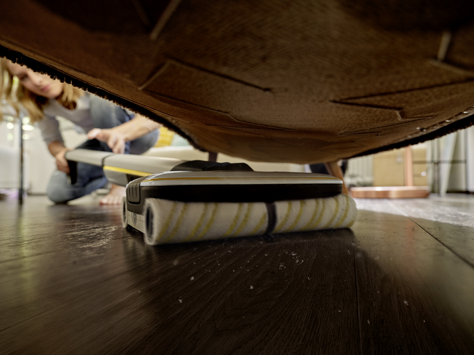 Kobieta odkurzająca pod dywanem za pomocą biało-żółtego odkurzacza.