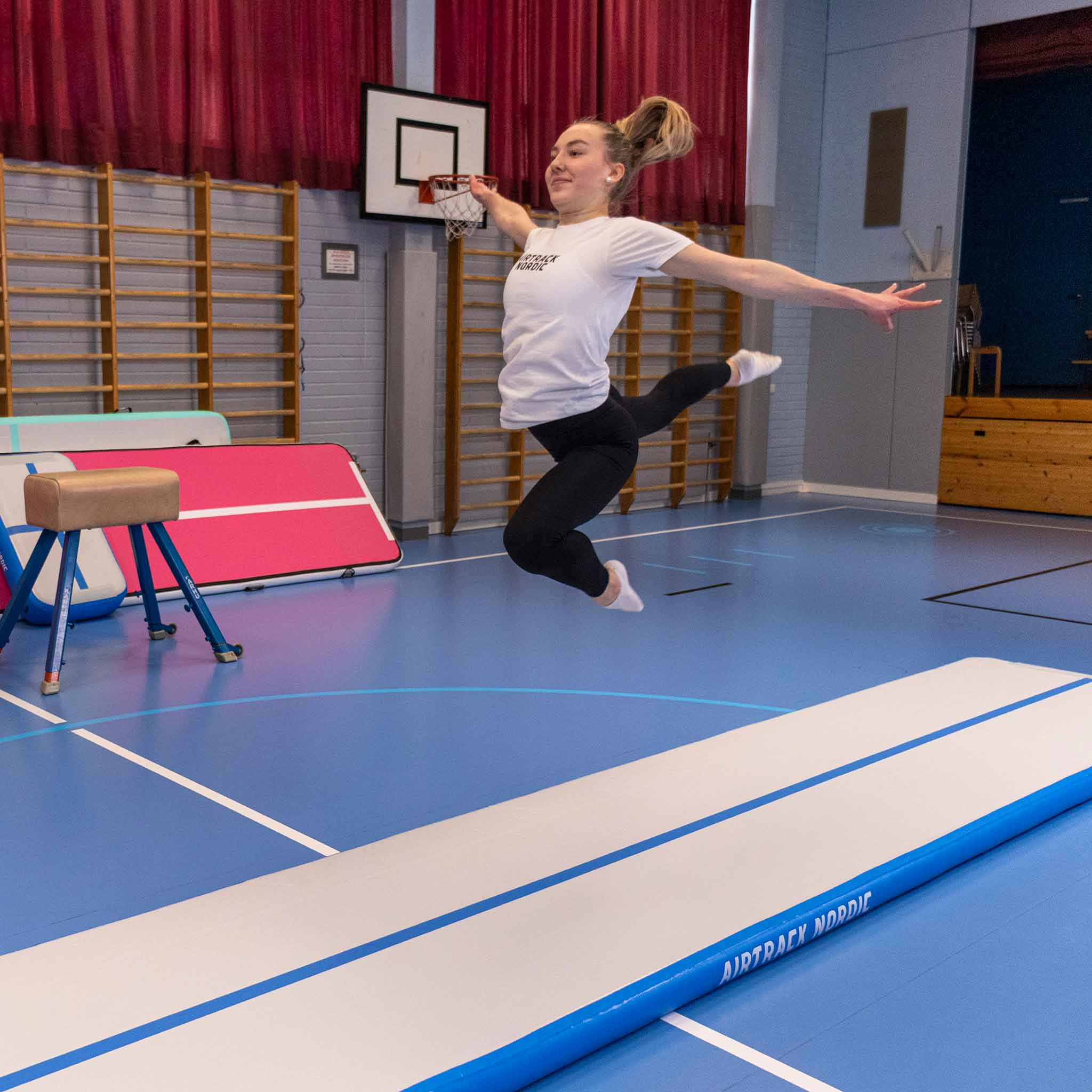 Młoda kobieta skacze na torze powietrznym w sali gimnastycznej, z koszem do koszykówki w tle.