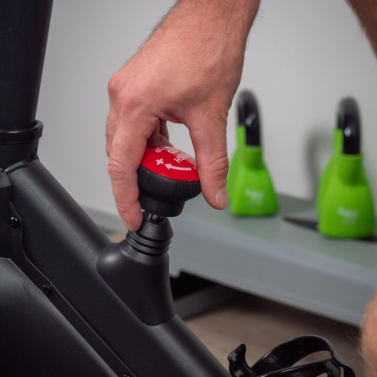 Dłoń reguluje czerwono-czarne pokrętło na rowerze stacjonarnym. Zielone ciężarki w tle.