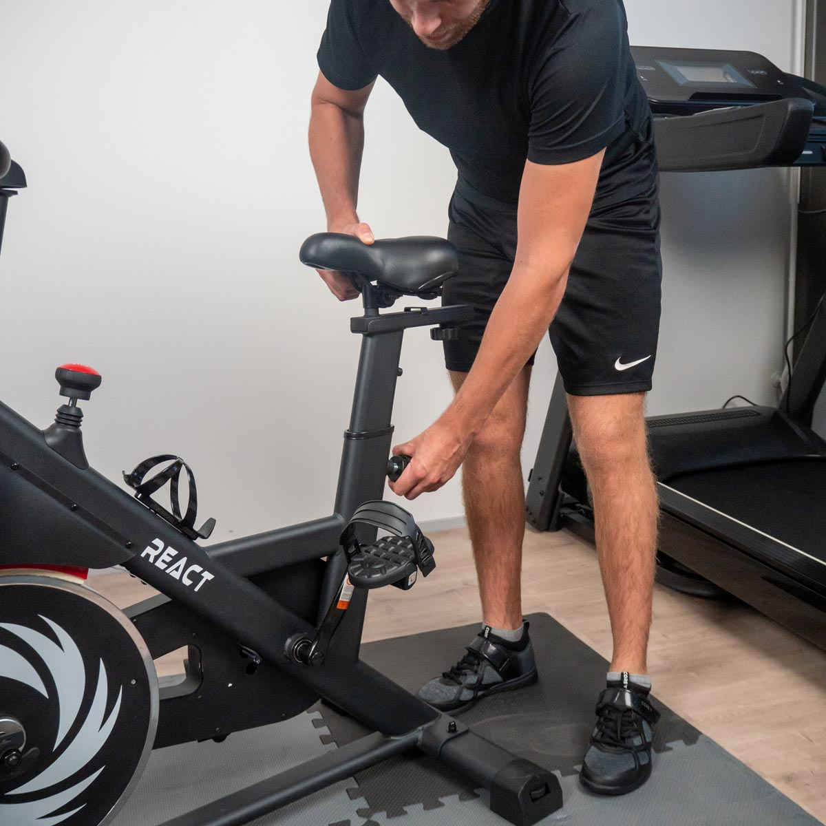 Mężczyzna reguluje siedzenie czarnego roweru stacjonarnego. Rower stoi na szarej macie.