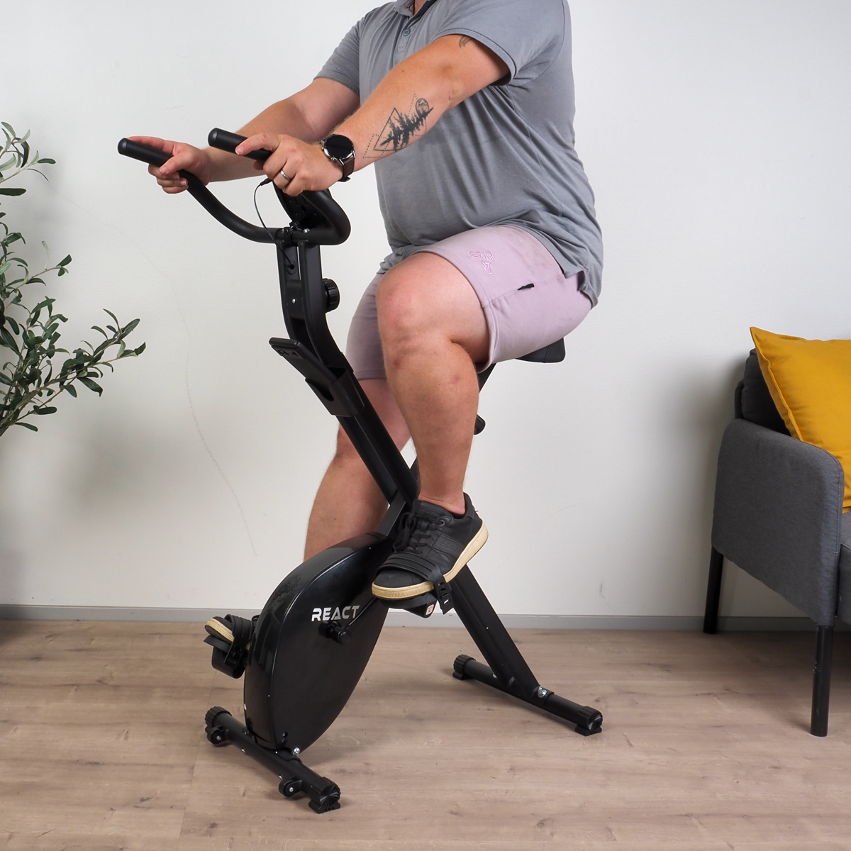 Mann fährt in einem Raum auf einem Trainingsrad. Er trägt ein graues Hemd, Shorts und hat ein Tattoo am Arm.