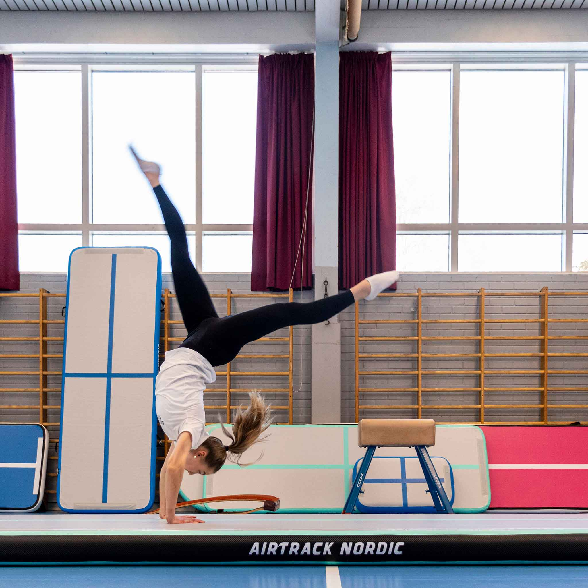 Gimnastyczka robiąca stanie na rękach na torze powietrznym. Czerwone zasłony i kosz do koszykówki w tle.