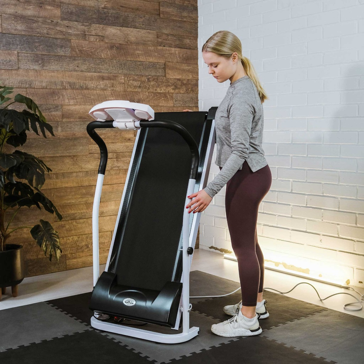 Kobieta regulująca bieżnię, stojąca obok ściany z cegieł i drewna. Szare i bordowe ubrania.