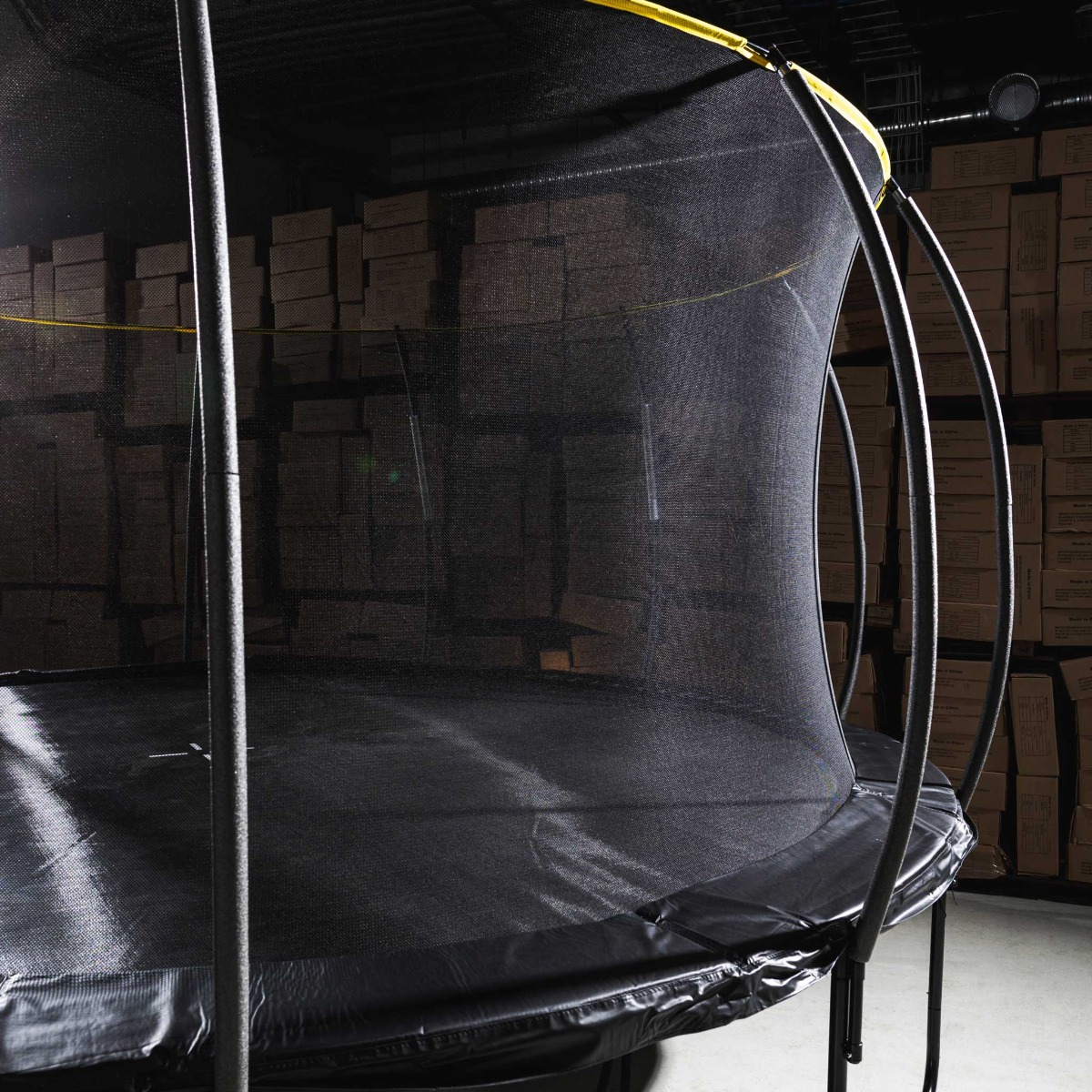 Ein schwarzes Trampolin mit Sicherheitsnetz in einer Lagerhalle. Kartons sind im Hintergrund gestapelt.