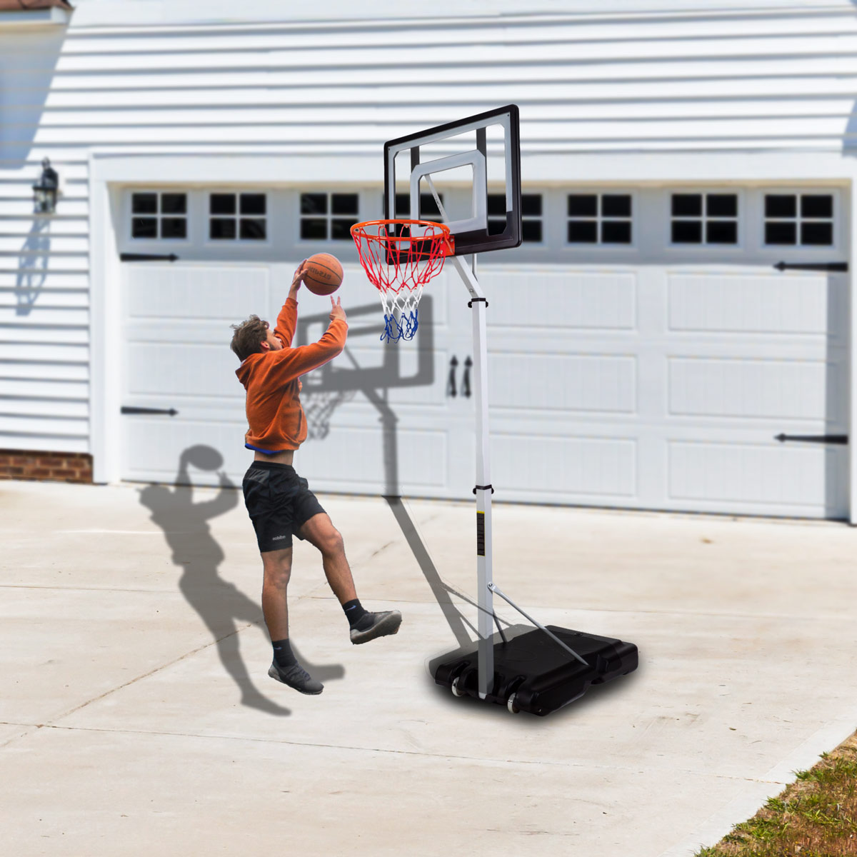 Chłopiec rzuca piłkę do kosza. Obręcz jest na czarnej podstawie, na podjeździe, z garażem.
