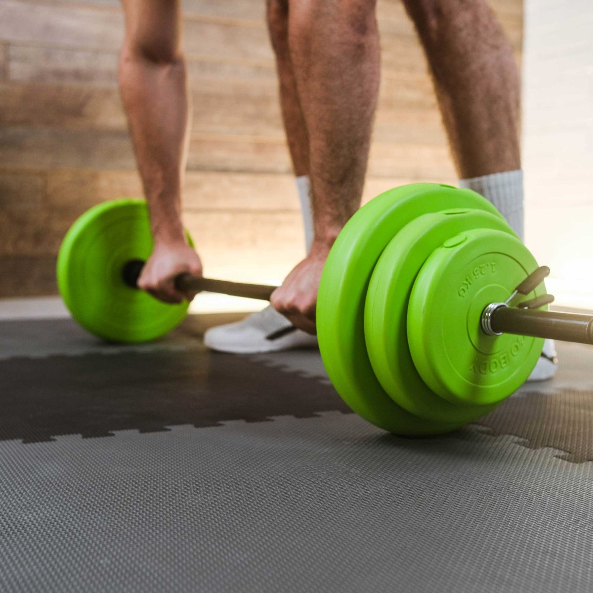 Mężczyzna podnosi sztangę z zielonymi ciężarami na szarej macie. Tło jest drewniane.