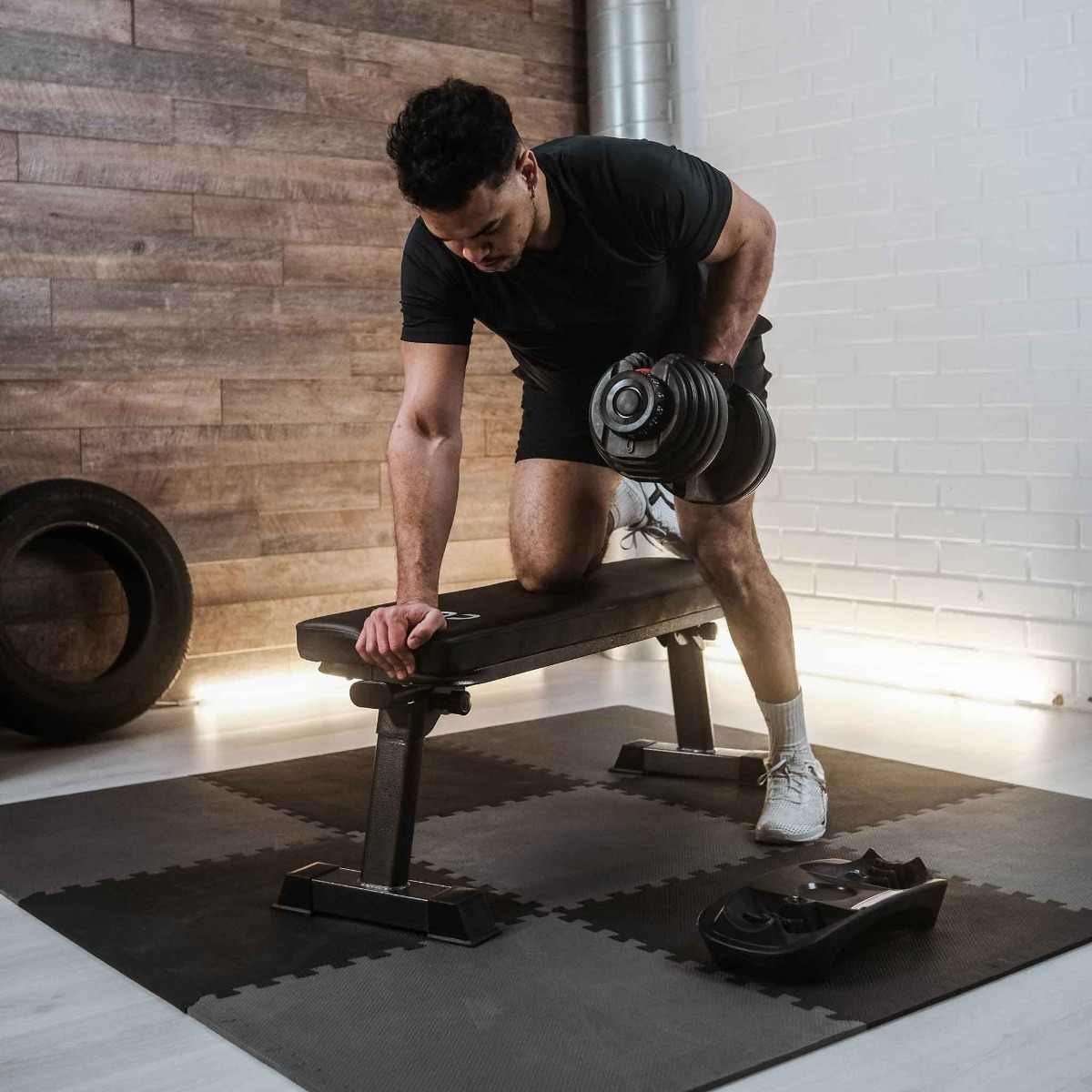 Ein Mann hebt eine Hantel, während er auf einer Hantelbank kniet. Er trägt Trainingskleidung in einem Fitnessstudio.