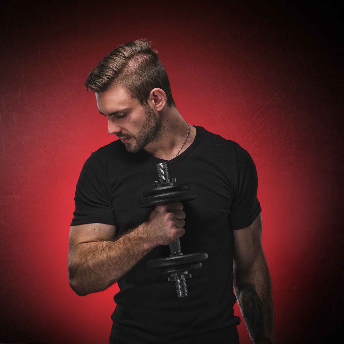 Mężczyzna w czarnej koszulce trzymający hantle, pozujący na czerwonym tle.