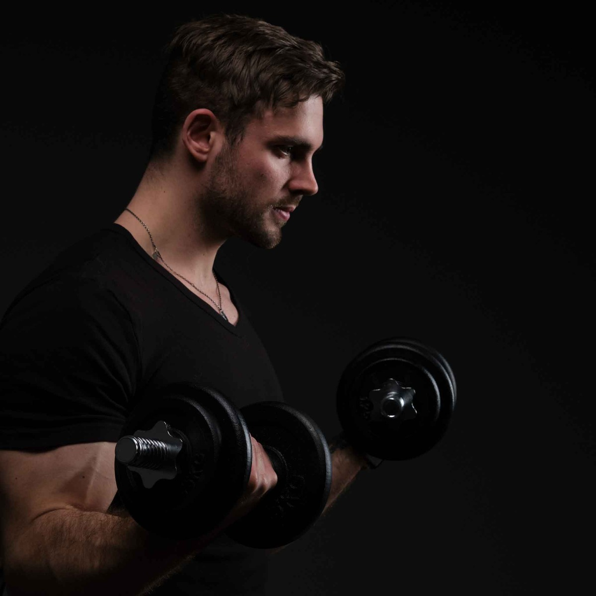 Mężczyzna podnosi ciężary, ciemne tło. Ma na sobie czarną koszulę, patrzy w dół.