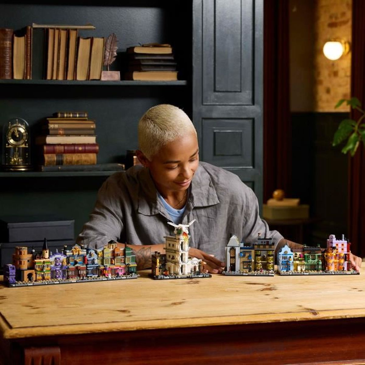 Osoba z blond włosami siedzi przy stole z budynkami Lego i półkami w tle.