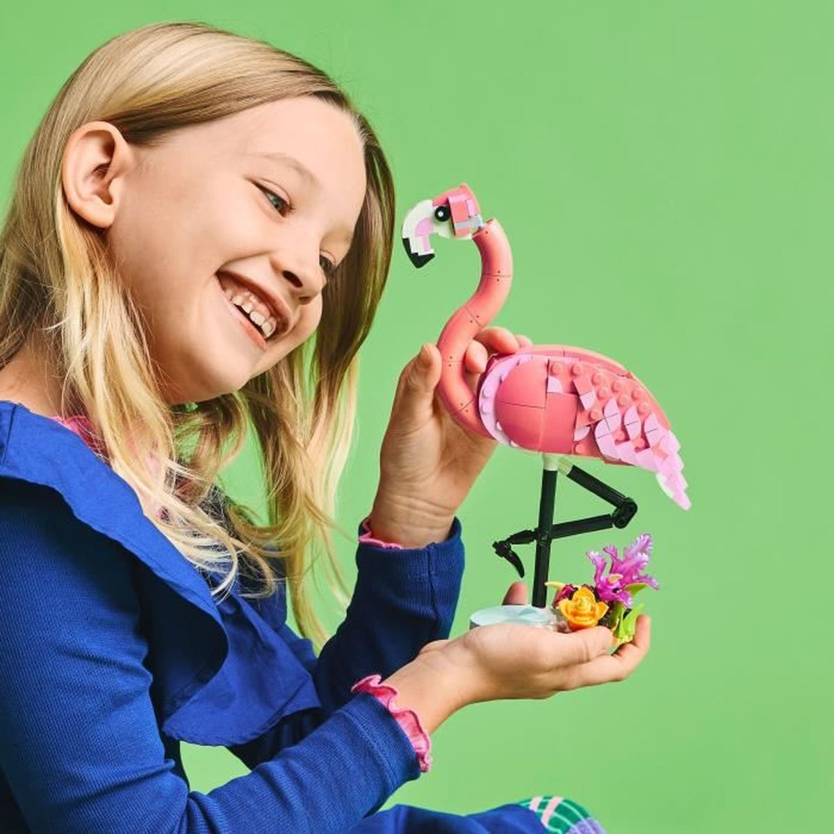 Dziewczynka trzymająca różowego flaminga Lego. Uśmiecha się. Zielone tło z kwiatami.