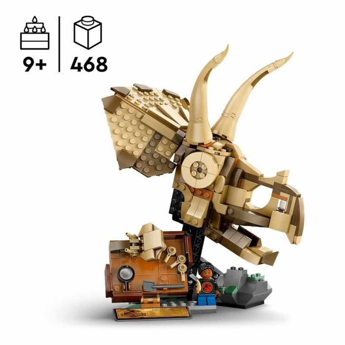 Una cabeza de Triceratops de Lego con cuernos grandes. Es marrón y beige. Una minifigura está al lado de una caja marrón y una roca.